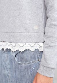 Ljusgrå sweatshirt med spetsdetalj i nederkant, med en diskret etikett, kombinerad med blå randiga denimbyxor. Mjuk textur och avslappnad passform.