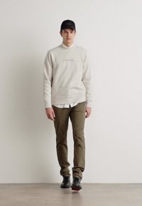 Lys grå hettegenser med liten tekst, hvit collared skjorte under, olivengrønne bukser og svart caps, iført med svarte sko.