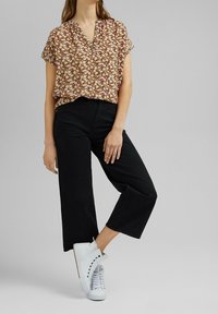 Donna che indossa una blusa floreale a maniche corte, pantaloni cropped neri a gamba larga e sneakers alte bianche su uno sfondo grigio neutro.