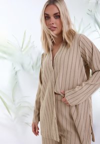 Camicia lunga beige e texturizzata con righe verticali, chiusura a bottoni e vestibilità rilassata, abbinata a pantaloni coordinati. Sullo sfondo si possono vedere delle morbide foglie verdi.