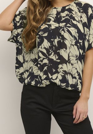 Femme portant un chemisier ample beige et noir à motif floral avec manches courtes et un pantalon noir, main posée sur la hanche.