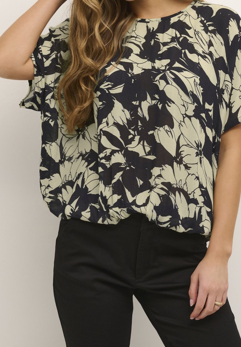 Femme portant un chemisier ample beige et noir à motif floral avec manches courtes et un pantalon noir, main posée sur la hanche.