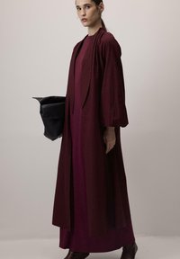 Longue robe d'extérieur bordeaux superposée à une robe cintrée de couleur similaire. Tissu doux avec une finition texturée, dotée d'un col et d'une ouverture sur le devant.