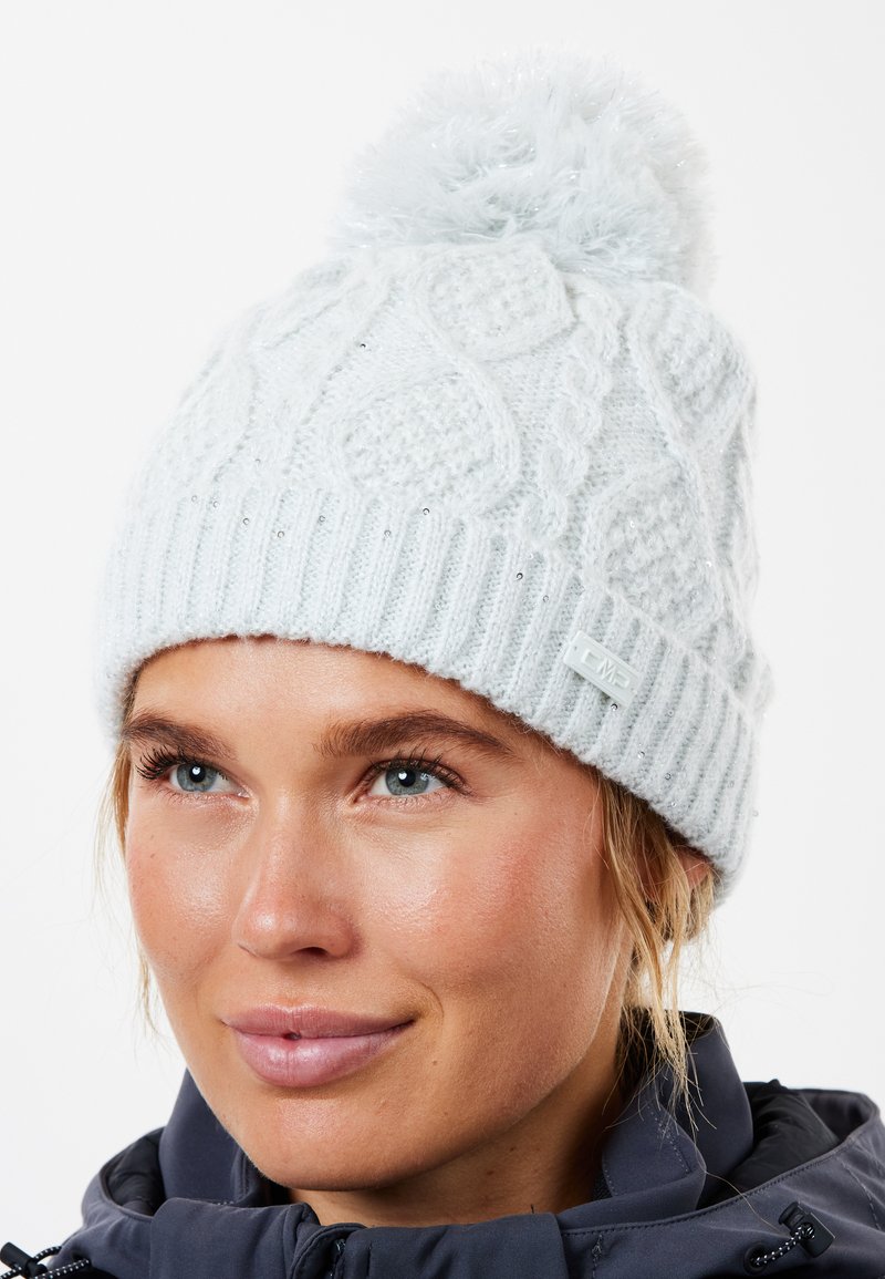 Lys grå strikket beanie med et struktureret mønster og en fluffy pompon på toppen; har et lille mærke på siden.