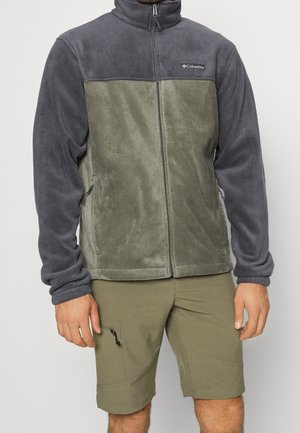 Mann trägt zweifarbige Columbia-Fleecejacke in Grau und Olivgrün mit olivgrünen Shorts, steht mit entspannt seitlich hängenden Händen.
