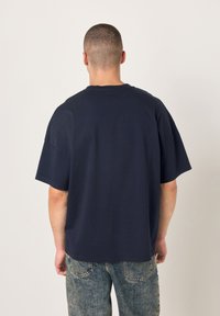 Bombažna majica mornarsko modre barve z ohlapnim krojem, kratkimi rokavi in okroglim izrezom. Pogled od zadaj prikazuje gladko teksturo in minimalne detajle.