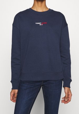 Persoon draagt een donkerblauwe trui met het "TOMMY JEANS"-logo in wit en rood tekst, gecentreerd op de borst, gecombineerd met donkerblauwe spijkerbroek.