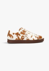 Zapatillas blancas con estampado de vaca marrón, que cuentan con una parte superior texturizada, cierre de cordones y suela de goma marrón con un diseño de calavera y huesos cruzados.