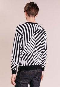 Gestreifter schwarzer und weißer Pullover mit geometrischen Mustern, langen Ärmeln und ribbeltem Saum, von hinten an einem Modell präsentiert.