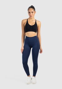 Svart sportbh med rynkning och justerbara axelband, kombinerad med högmidjade marinblå tights. Modellen har på sig vita träningsskor.