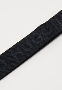 Ceinture tissée noire étroite avec texte en relief, présentant une texture lisse et des bords nets, conçue pour un usage décontracté.