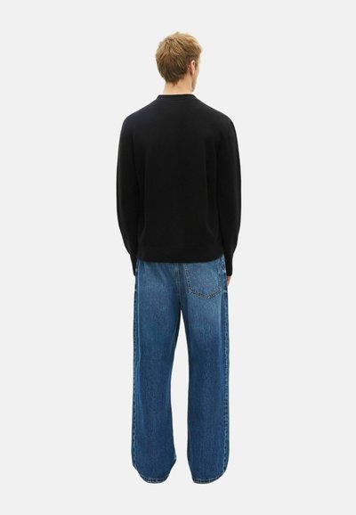 Mand iført en sort langærmet sweater og løse blå jeans, stående med ryggen til mod en ensfarvet baggrund.