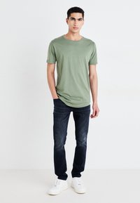 Camiseta verde de manga corta, de tela suave, con cuello redondo; combinada con jeans de mezclilla oscura y zapatillas blancas; un atuendo sencillo y casual.