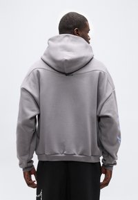 Mann trägt einen grauen übergroßen Hoodie mit großer Kapuze, steht mit dem Rücken zur Kamera, zeigt Details auf dem Rücken sowie schwarze Hosen mit weißem Muster.