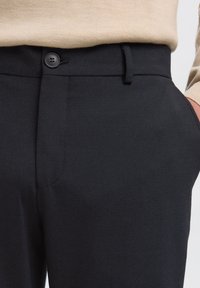 Primo piano della vita di pantaloni neri con bottone e passante per cintura, maglione beige infilato e mano nella tasca destra.