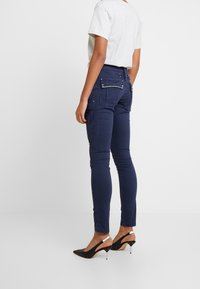 Granatowe spodnie cargo o dopasowanym kroju, z ozdobnymi ćwiekami na tylnej kieszeni i gładką teksturą. Noszone z czarnymi, wysokimi szpilkami.