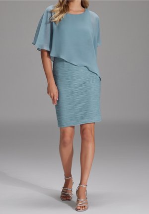 Frau trägt ein hellblaues, mehrlagiges Kleid mit Flatterärmeln und silberne, gestreifte Sandaletten mit Absatz, steht vor einem schlichten grauen Hintergrund.