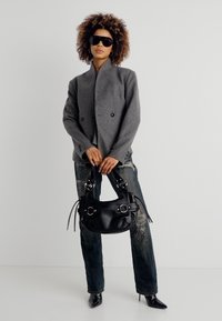 Γυναίκα με κατσαρά μαλλιά που φοράει μεγάλα μαύρα γυαλιά ηλίου, γκρι σακάκι με διπλό πέτο, σκισμένο σκούρο τζιν, μαύρες μυτερές γόβες, κρατώντας μαύρη τσάντα.