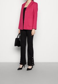 Blazer sartorial rosa con due tasche frontali, indossato sopra un top nero, abbinato a pantaloni neri a palazzo con dettagli di bottoni. Tacchi alti neri e borsa.