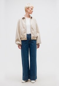 Άτομο με κοντά ξανθά μαλλιά που φοράει μπεζ μπουφάν με φερμουάρ, λευκό τοπ, μπλε τζιν με φαρδιά μπατζάκια και λευκά αθλητικά παπούτσια, κοιτώντας πλάγια.