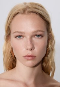 Modell med långt blont hår och klar hy, som bär geometriska guldsmycken. Naturlig makeup med ljusrosa inslag och diskreta fräknar.