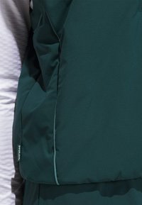 Tummanvihreä liivi pehmeästä, teksturoidusta kankaasta. Varustettu sileällä vetoketjulla ja sivulla logotarralla. Paneelimuotoilu, jossa on kontrastivaaleat saumat.