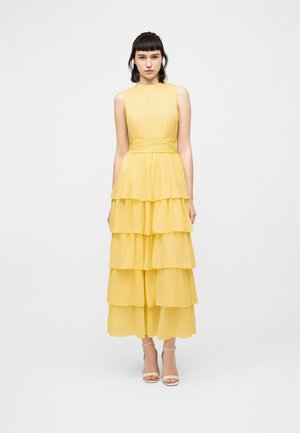 Mujer de pie, vistiendo un vestido maxi amarillo sin mangas con volantes y cinturilla ajustada, y tacones blancos abiertos, sobre un fondo blanco liso.