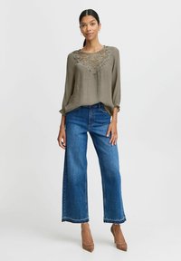 Blusa verde oliva con dettagli in pizzo intorno al collo, abbinata a jeans a gamba larga blu e scarpe con tacco color nocciola.