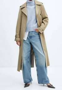 Beiger Trenchcoat über einem hellblauen gerippten Rollkragenpullover, kombiniert mit weiten hellblauen Jeans und schwarzen spitzen Absatzschuhen. Glatte Texturen.