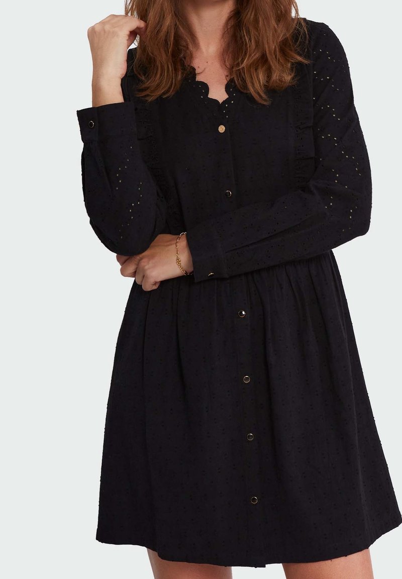 Mujer vestida con un vestido negro de manga larga y botones en la parte delantera, con detalles de ojales, de pie con un brazo cruzado y la otra mano cerca de su rostro.