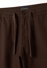 Pantaloni marroni a spina di pesce con chiusura a bottone e cordoncino. Presentano un'etichetta contrassegnata "PIOMBO" su uno sfondo blu scuro.
