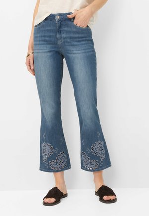 Kvinde iført blå korte flared jeans med broderede mønstre nær kanterne, hvid top og sorte sandaler med kors.