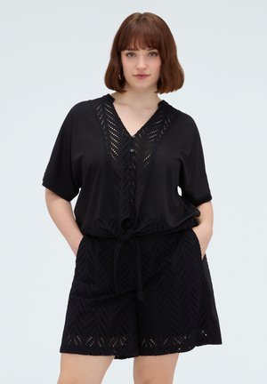 Femme aux cheveux courts bruns portant un haut noir boutonné à motif œillet et un short assorti, les mains dans les poches.