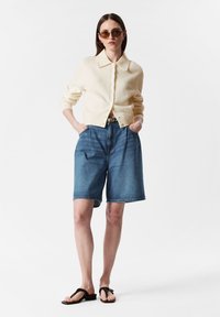 Cremfarbener Strickcardigan mit Kragen, Knopfleiste, kombiniert mit blauen Jeansshorts und schwarzen Flip-Flops. Das Model trägt braune Sonnenbrillen.