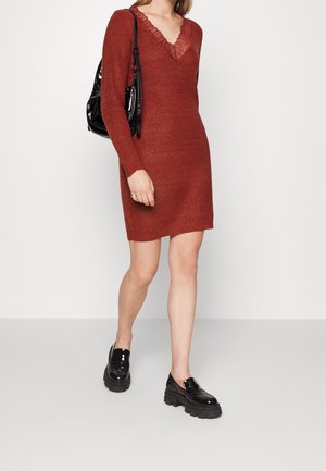 Robe tricotée couleur rouille avec une texture côtelée, des manches longues et un col en V bordé de dentelle. Associée à des mocassins en cuir verni noirs et un sac noir.
