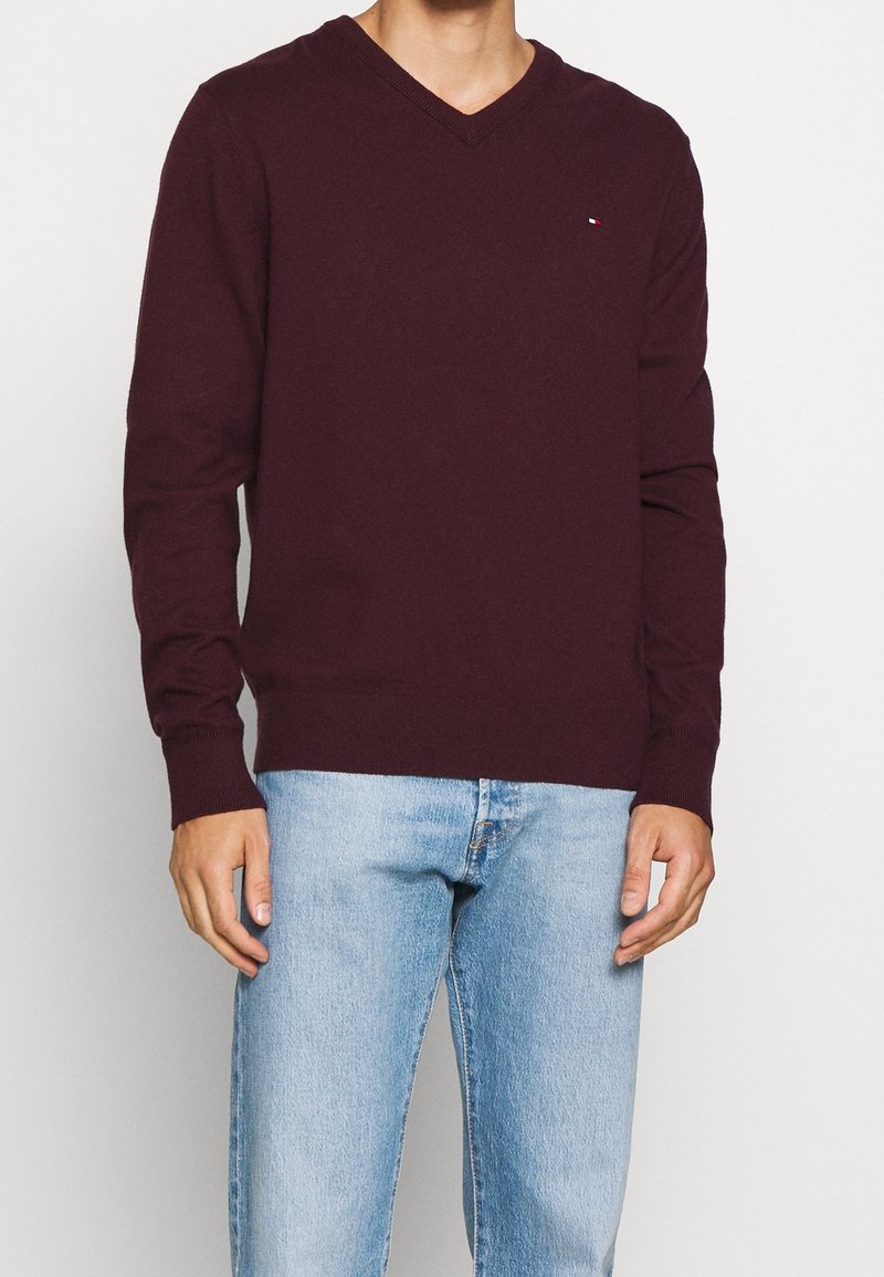 Homme portant un pull à col en V bordeaux foncé avec un petit logo sur la poitrine, associé à un jean en denim bleu clair sur un fond uni.