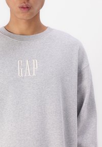 Szürke kapucnis pulóver, pamut keverékből készült, kerek nyakkivágással és laza szabással. A "GAP" logó hímzett, világosabb árnyalatban.