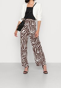 Ruskeat ja valkoiset seepra-kuosiset leveälahkeiset housut yhdistettynä mustaan ilmanhihaiseseen paitaansa ja valkoiseen lyhythihaiseen takkiin, asusteena musta käsilaukku.