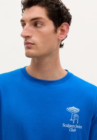 Blå bomulds t-shirt med rund hals, prydet med en hvid broderet grafik af en strandstol og parasol med teksten "Scalpers Swim Club".