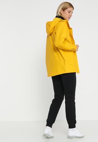 Geel waterdichte jas met een capuchon, middellange ontwerp, zijzakken, gecombineerd met zwarte broeken en witte sneakers. Gladde textuur.