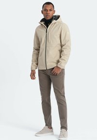 Beige Zip-Jacke mit Kapuze, strukturiertem Stoff, Fronttaschen und elastischen Bündchen. Kombiniert mit dunkelbraunen Hosen und hellen Sneakers.