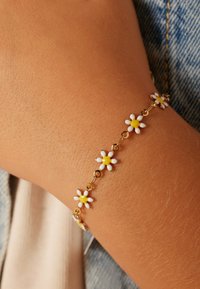 Gouden kettingarmband met kleine witte bloemen met gele centra, tegen een gebruinde huidskleur, die een delicate uitstraling toont.