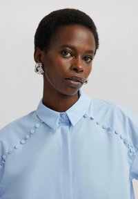 Camisa azul claro con cuello y detalles decorativos de volantes en las costuras de los hombros. Tejido suave con una silueta estructurada.