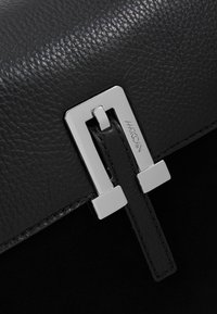 Gros plan sur le rabat en cuir noir texturé d'un sac avec une fermeture métallique rectangulaire argentée et une sangle en cuir passant à travers celle-ci.