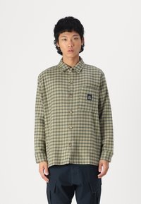 Green and beige checkered shirt with a button-up front, collar, long sleeves, and a chest pocket featuring a branded patch.