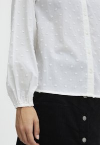 Weißes Blusenshirt mit strukturierten, erhabenen Punkten, Puffärmel und Knopfverschluss. Getragen mit einem schwarzen Cordrock mit silbernen Knöpfen.