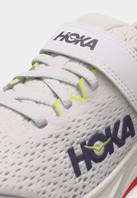 Κοντινό πλάνο λευκού παπουτσιού τρεξίματος Hoka με ύφασμα από δίχτυ, κορδόνια, λουράκι και λεπτομέρειες λογότυπου σε μοβ και νέον κίτρινο χρώμα.