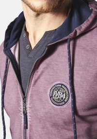 Purple zip-up hoodie featuring a navy hood, front zipper, and a round logo patch. Underneath, a dark gray V-neck shirt is visible.