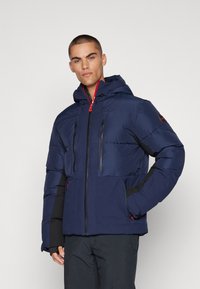 Navy puffer jacket featuring a hood, two zippered chest pockets, and contrasting black sleeve accents with red zipper details.