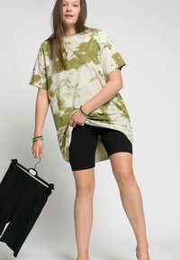 Oversized tie-dye t-shirt in beige and olive green with short sleeves, paired with black bike shorts. Model holds a black skirt on a hanger.
